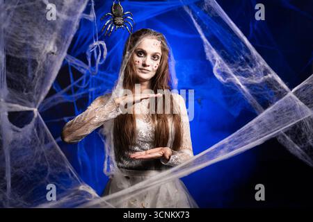 Unheimliche Frau in weißem schmutzigem Kleid mit schrecklichem Halloween Zombie Make-up, die große Geste zeigt, mit ihren Fingern, die auf blauem Hintergrund stehen Stockfoto