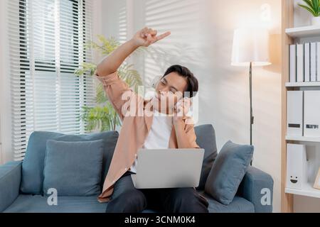Ein fröhlicher junger Mann sitzt auf einem Sofa und singt mit Leidenschaft, während er einen Laptop hält. Seine fröhlichen Gesten und sein heller Ausdruck spiegeln den Spaß der Musik wider Stockfoto