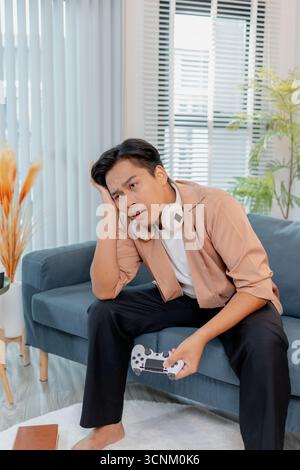 Ein junger Mann sitzt auf einem Sofa mit einem Game-Controller, voll konzentriert und frustriert, während er ein Videospiel spielt. Seine intensive Reaktion ist hochgradig Stockfoto