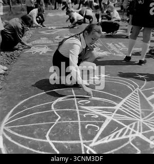 Ein stimmungsvolles Schwarzweiß-Foto fängt eine Kreidezeichnung auf Asphaltkonkurrenz in Sloviansk in den 1970er Jahren ein; eine Gruppe sowjetischer Pioniere in Schuluniformen erstellt enthusiastisch große patriotische Zeichnungen zu den Themen Raum, Frieden und Heldentum unter der Aufsicht von Lehrern, was ein klassisches Beispiel für kollektive Kreativität und ideologische Erziehung von Kindern in der UdSSR ist Stockfoto
