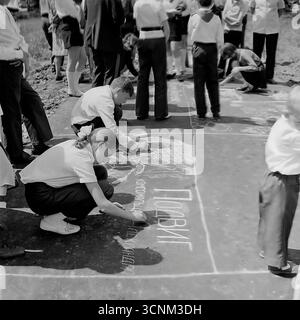 Ein stimmungsvolles Schwarzweiß-Foto fängt eine Kreidezeichnung auf Asphaltkonkurrenz in Sloviansk in den 1970er Jahren ein; eine Gruppe sowjetischer Pioniere in Schuluniformen erstellt enthusiastisch große patriotische Zeichnungen zu den Themen Raum, Frieden und Heldentum unter der Aufsicht von Lehrern, was ein klassisches Beispiel für kollektive Kreativität und ideologische Erziehung von Kindern in der UdSSR ist Stockfoto