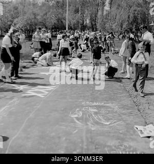 Ein stimmungsvolles Schwarzweiß-Foto fängt eine Kreidezeichnung auf Asphaltkonkurrenz in Sloviansk in den 1970er Jahren ein; eine Gruppe sowjetischer Pioniere in Schuluniformen erstellt enthusiastisch große patriotische Zeichnungen zu den Themen Raum, Frieden und Heldentum unter der Aufsicht von Lehrern, was ein klassisches Beispiel für kollektive Kreativität und ideologische Erziehung von Kindern in der UdSSR ist Stockfoto
