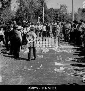 Ein stimmungsvolles Schwarzweiß-Foto fängt eine Kreidezeichnung auf Asphaltkonkurrenz in Sloviansk in den 1970er Jahren ein; eine Gruppe sowjetischer Pioniere in Schuluniformen erstellt enthusiastisch große patriotische Zeichnungen zu den Themen Raum, Frieden und Heldentum unter der Aufsicht von Lehrern, was ein klassisches Beispiel für kollektive Kreativität und ideologische Erziehung von Kindern in der UdSSR ist Stockfoto