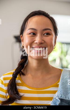 Frau lächelt, während sie die Jeansjacke über die Schulter in einem sanft beleuchteten Wohnzimmer hält Stockfoto