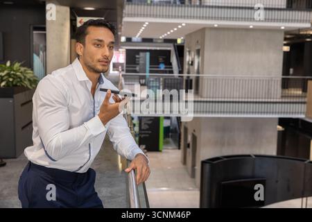 Mann im Anzug, der sich auf das Geländer im Atrium des Büros lehnt und das Smartphone an den Mund hält, Kopierraum Stockfoto