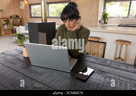 Asiatische Frau Mitte dreißiger, die auf Laptop in offener Kabine Küche mit Smartphone, Becher tippt Stockfoto