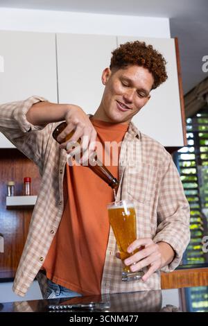 Rothaariger Mann, der Bier aus der Flasche in Pilsner-Glas in der Küche mit Metallöffner gießt Stockfoto