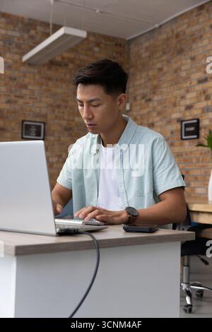 Asiatischer Mann, der an einem Laptop am Holzschreibtisch im Backsteinbüro mit Smartphone-Kabel arbeitet Stockfoto