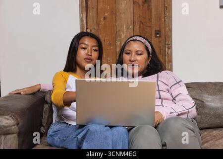 Vielfältige Mutter und Tochter sitzen dicht auf Ledercouch im Wohnzimmer mit silbernem Laptop Stockfoto