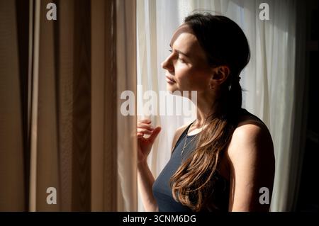 Eine junge Frau sitzt drinnen und blickt nachdenklich aus dem Fenster, während warmes Sonnenlicht auf sie fällt, was eine friedliche und gemütliche Atmosphäre schafft Stockfoto