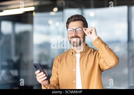 Junger Geschäftsmann in Smart-Casual-Kleidung lächelt selbstbewusst, während er die Brille anpasst und ein Smartphone hält, um digitale Kommunikation, Netzwerke und modernen Erfolg am Arbeitsplatz zu vermitteln Stockfoto