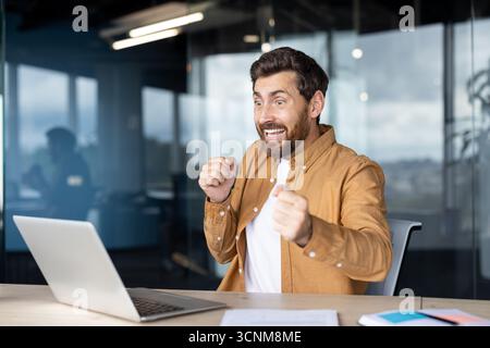 Glücklicher bärtiger Mann, der Triumph und Sieg im Amt feiert, auf den Laptop-Bildschirm blickt und Fäuste geballt, sich über gute Nachrichten oder erreichte Ziele freut Stockfoto
