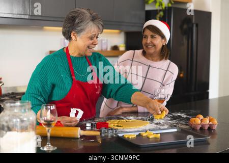 Seniorenmutter und Erwachsene Tochter schneiden Weihnachtskeksteig auf der Theke mit Nudelnadel, Schneidemaschinen Stockfoto