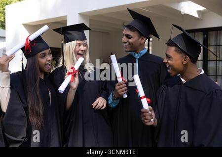 Verschiedene Absolventen feiern unter der weißen Veranda mit Diplomen mit roten Bändern in Kappen und Gewändern Stockfoto