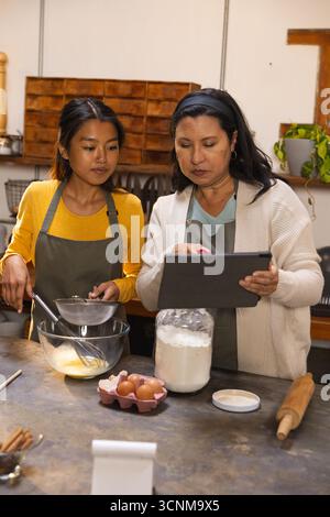 Vielfältige Mutter und Tochter in Schürzen Mehl in Schüssel gießen, Rezept auf Tablette lesen in der Küche Stockfoto
