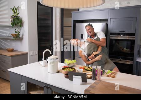 Seniorenpaar, das Schürzen trägt, tanzt auf der Kücheninsel in der Küche und bereitet Salat vor Stockfoto