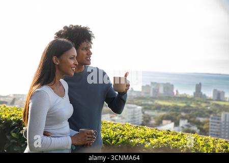 Verschiedene Paare stehen auf der Dachterrasse an der Hecke mit Blick auf die Küstenstadt und halten Tassen in der Hand Stockfoto