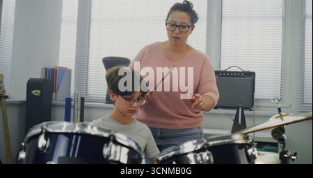Der Grundschuljunge übt Sinn für Rhythmus, spielt Schlagzeug in der Musikklasse und träumt davon, Schlagzeuger zu werden. Eine Lehrerin, die talentierten Jungen während des Percussion-Lessons zum Trommelspielen erzieht. Musikalische Kunst Stockfoto