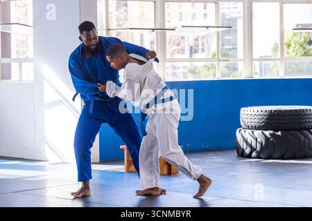 Verschiedene männliche Trainingspartner in blau und weiß gis, die auf gepolstertem Studioboden einen Überwurf durchführen Stockfoto