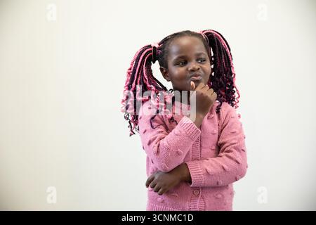 afroamerikanisches Mädchen in rosa Pullover, lehnt sich an eine schlichte Wand und liegt die Hand auf dem Kinn Stockfoto