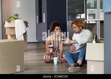 Verschiedene Paare sitzen in einem neuen Wohnzimmer zwischen Boxen, die Smartphone zeigen, Kaffee halten und Lampe Stockfoto