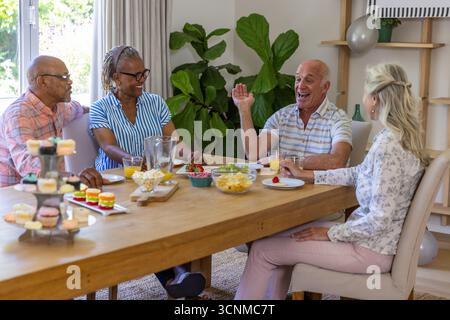 Verschiedene Seniorenfreunde unterhalten sich zu Hause am Esstisch mit Cupcake-Stand, Orangensaft Stockfoto