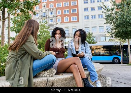 Drei junge Frauen sitzen auf einer niedrigen Wand, reden und benutzen ihre Smartphones in einer städtischen Umgebung Stockfoto