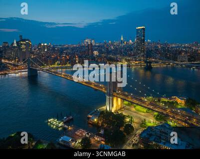 Die beleuchtete Brooklyn Bridge aus der Vogelperspektive erstreckt sich über den East River und verbindet die glitzernde Skyline von Manhattan unter der verblassenden Dämmerung in Manhattan, New York, USA. Stockfoto