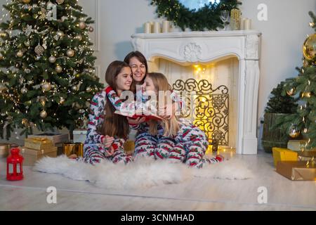 Glückliche Familie liest zusammen am Weihnachtsmorgen. Mutter und Töchter im passenden Pyjama kuscheln und lesen ein Buch am Weihnachtsbaum und Stockfoto