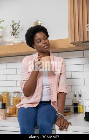 MittelErwachsene afroamerikanische Frau, die auf der Arbeitsplatte in der Küche sitzt und einen Keramikbecher hält Stockfoto