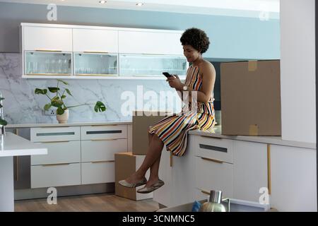 afroamerikanerin im Kleid, die auf der Küchentheke sitzt und das Smartphone mit Kisten überprüft Stockfoto
