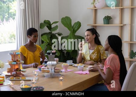 Verschiedene Freundinnen sitzen zu Hause um hölzernen Esstisch, essen Cupcakes und plaudern Stockfoto