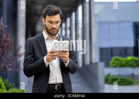 Geschäftsmann steht im Freien, konzentriert sich auf ein digitales Tablet, verwaltet geschäftliche Aufgaben, stellt eine Verbindung zur Technologie her, repräsentiert Mobilität und professionelles c Stockfoto