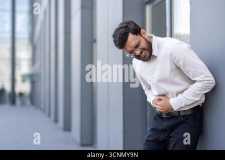 Ein Mann, der seinen Magen wegen Verdauungsstörungen, Lebensmittelvergiftungen oder Krämpfen unter Schmerzen hält und sich krank fühlt, während er sich an eine moderne Gebäudewand lehnt Stockfoto