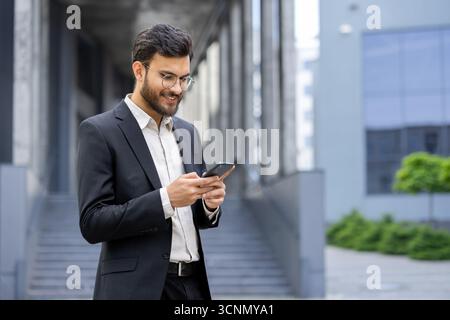 Junger Geschäftsmann mit Brille und Anzug, der draußen vor einem städtischen Gebäude steht und lächelt, während er das Mobiltelefon für Kommunikation und Geschäftsleute benutzt Stockfoto