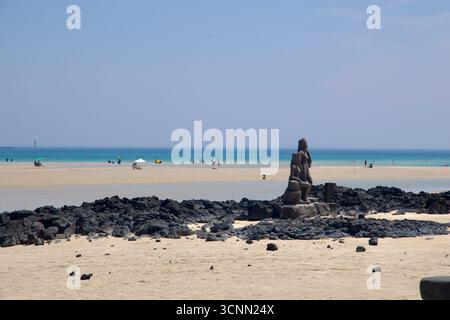 Zwei Statuen, die Jejus haenyeo-Taucher darstellen, stehen zwischen schwarzen Basaltvorsprüngen auf der Sandbank am Pyoseon Beach, mit Badegästen, die über den ganzen wi verstreut sind Stockfoto