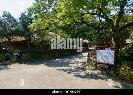 Ein Schild und ein kurviger Pfad führen zwischen Steinmauern und strohgedeckten Hütten in der von Laubbäumen beschatteten Bergzone des Jeju Folk Village. Stockfoto