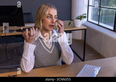 Blonde Frau, die das Smartphone in der Nähe des Mundes hält und sich mit der Tastatur am Schreibtisch unter dem Auge berührt Stockfoto