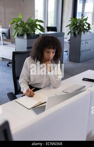 afroamerikanische Frau, die auf Laptop schreibt und mit Bleistift in Notizbuch schreibt, am Schreibtisch im Büro Stockfoto