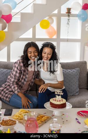 Vielfältige Mutter und Tochter im Teenageralter sitzen auf dem Sofa bei der Treppe und feiern Geburtstag mit Kuchenballons Stockfoto