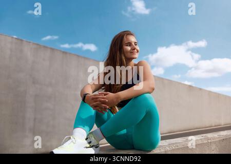 Athletische Frau mit langen Haaren in Trainingskleidung sitzt auf einer Betonbarriere und genießt die Natur und verkörpert einen entspannten, aber aktiven Lebensstil Stockfoto
