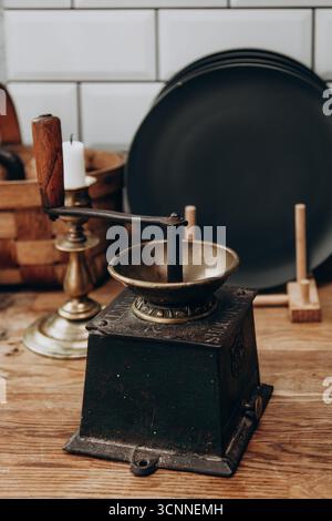 Eine Vintage Kaffeemühle sitzt auf einem rustikalen Holztisch, umgeben von einer Kerze, Tellern und Küchenutensilien. Die Szene fängt eine warme, gemütliche Atmosphäre ein Stockfoto