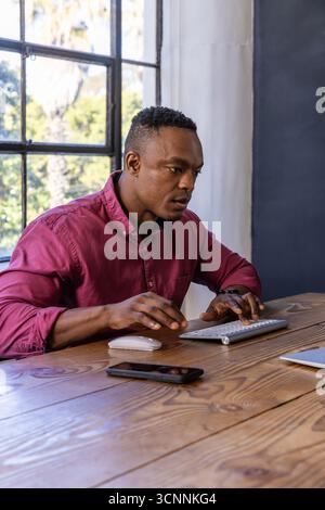 afroamerikanischer Mann, der drahtlose Tastatur und Maus auf Laptop benutzt und Smartphone am Tisch überprüft Stockfoto