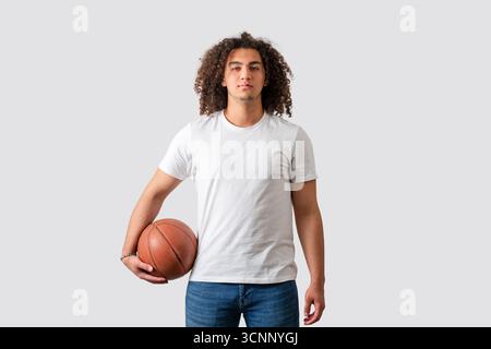 Selbstbewusster junger Mann, der Basketball in lässigem Outfit auf weißem Hintergrund hält. Hochwertige Fotos Stockfoto