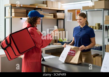 Kaukasische junge Erwachsene Frau in blauem Poloshirt, die Essensbeutel an kaukasische junge Erwachsene weibliche Kurierin mit Mütze und rotem isoliertem Rucksack überreicht, beide lächeln im Lagerhaus Stockfoto