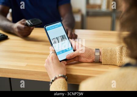 Kaukasische junge Erwachsene Frau, die ein Smartphone hält, das Barcode anzeigt, während Black man einen Scanner mit handgehaltenem Barcode-Leser über eine Holztheke in moderner Einzelhandels- oder Versandumgebung scannt Stockfoto