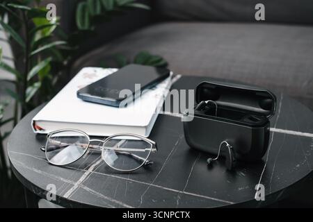 Moderne digitale Hörgeräte mit Ladetasche auf schwarzem Marmortisch, Brille, Smartphone und Notebook. Täglicher Zugang für Profis Stockfoto