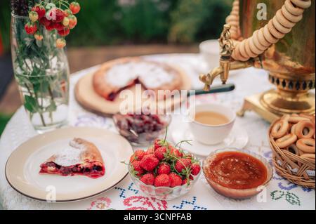 Eine rustikale Teeumgebung mit goldenem Samovar, frischen Erdbeeren, Marmelade, Kuchen und Bagels, die auf einer gestickten Tischdecke im Freien angeordnet sind. Stockfoto