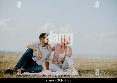 Ein Paar sitzt zusammen auf einer Decke auf einem Feld unter klarem Himmel. Sie lächeln und schauen sich an und genießen einen friedlichen und sonnigen Tag im Freien Stockfoto