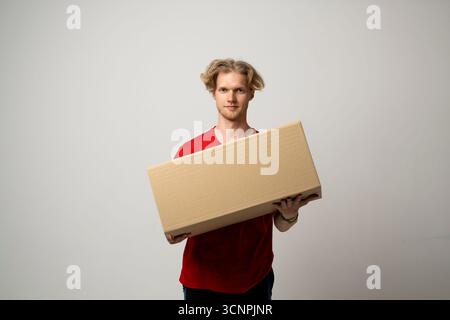 Junger Mann in rotem T-Shirt, der Karton auf neutralem Atelierhintergrund hält Stockfoto